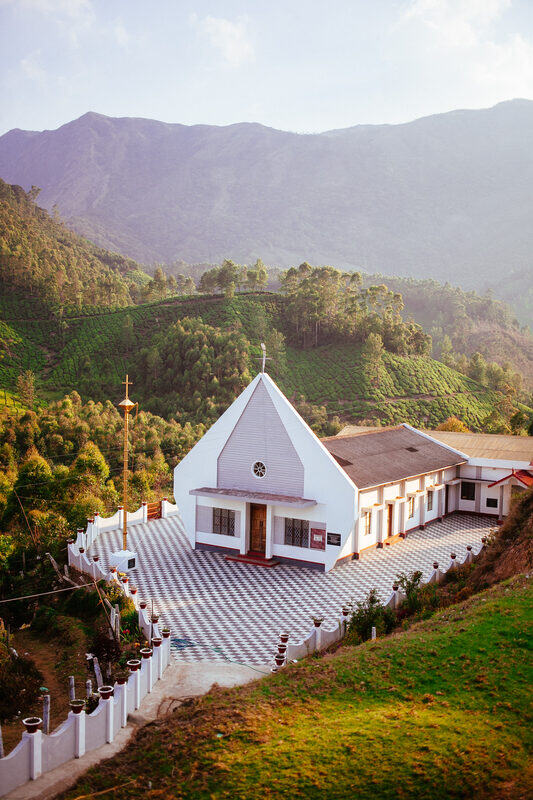 A white Catholic chapel sits below tea-covered mountains near Munnar Top Station, reflecting the Catholic and Portuguese layers in the region. print option