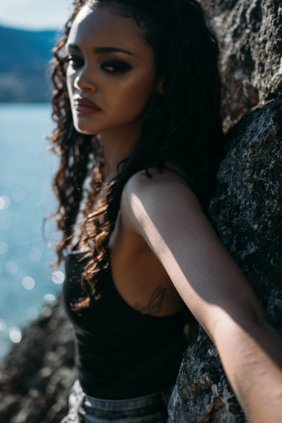 Alex by the Lake portraits story frame 3: Okanagan Lake portraits in Kelowna with black summer styling, rock, water, wind, direct expression and a cinematic beauty-portrait edge.