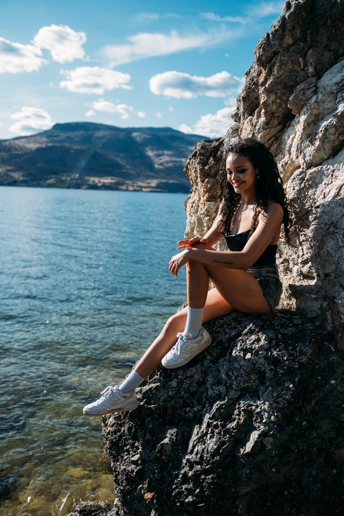 Alex by the Lake portraits story frame 4: Okanagan Lake portraits in Kelowna with black summer styling, rock, water, wind, direct expression and a cinematic beauty-portrait edge.