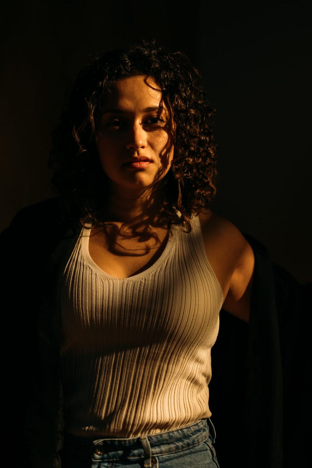 Marina in Warm Window Light portraits story frame 3: Marina in warm indoor light, with amber shadow, denim, black robe, green styling and an intimate editorial portrait mood.