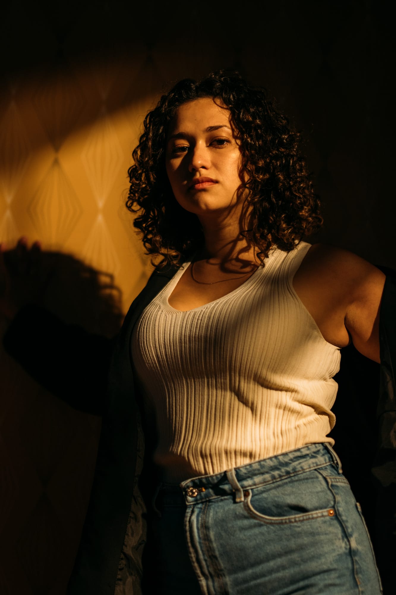 Marina in Warm Window Light portraits story frame 9: Marina in warm indoor light, with amber shadow, denim, black robe, green styling and an intimate editorial portrait mood.