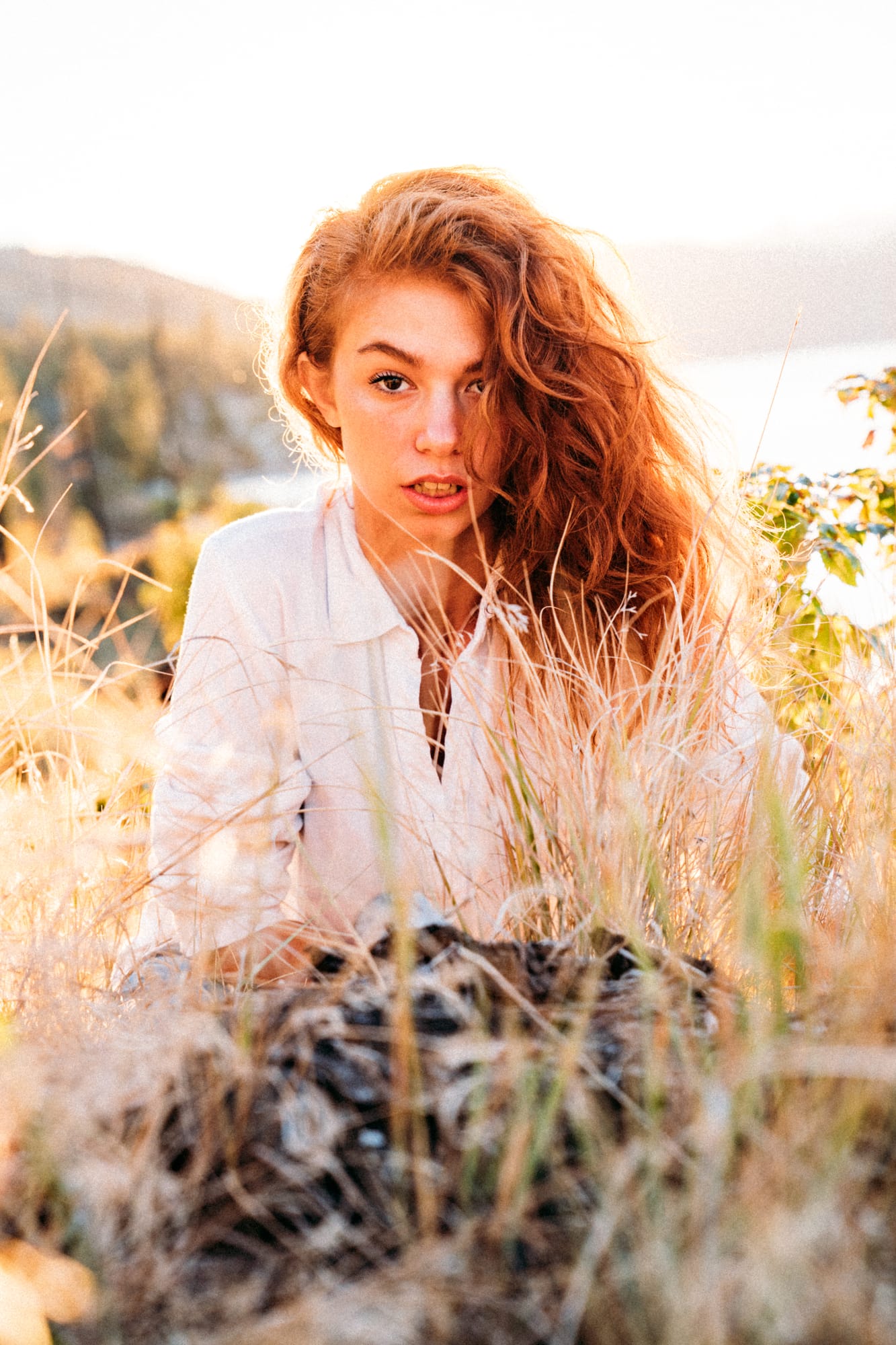 Mickie portraits story frame 6: A Kelowna portrait session for Mickie moving from golden grass and warm hillside light into lake-edge rocks, wind, white shirt styling and close editorial portraits.