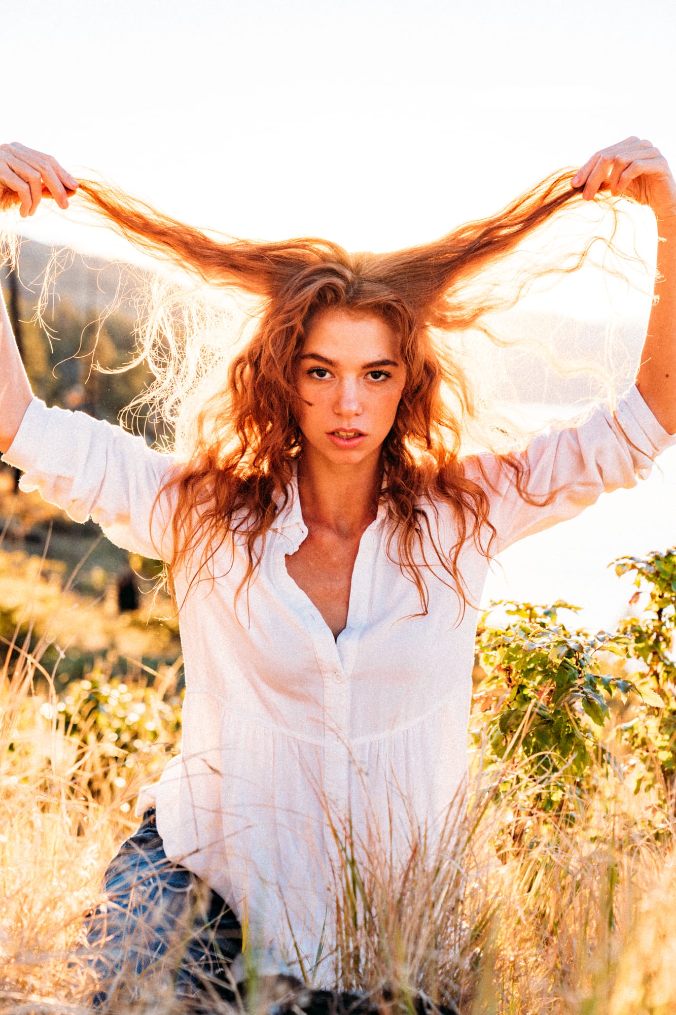 Mickie portraits story frame 7: A Kelowna portrait session for Mickie moving from golden grass and warm hillside light into lake-edge rocks, wind, white shirt styling and close editorial portraits.