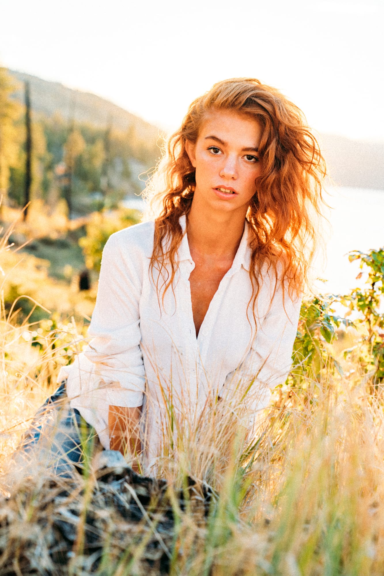 Mickie portraits story frame 8: A Kelowna portrait session for Mickie moving from golden grass and warm hillside light into lake-edge rocks, wind, white shirt styling and close editorial portraits.