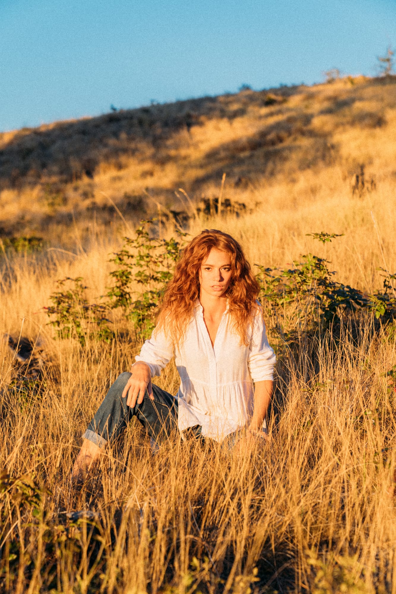Mickie portraits story frame 9: A Kelowna portrait session for Mickie moving from golden grass and warm hillside light into lake-edge rocks, wind, white shirt styling and close editorial portraits.