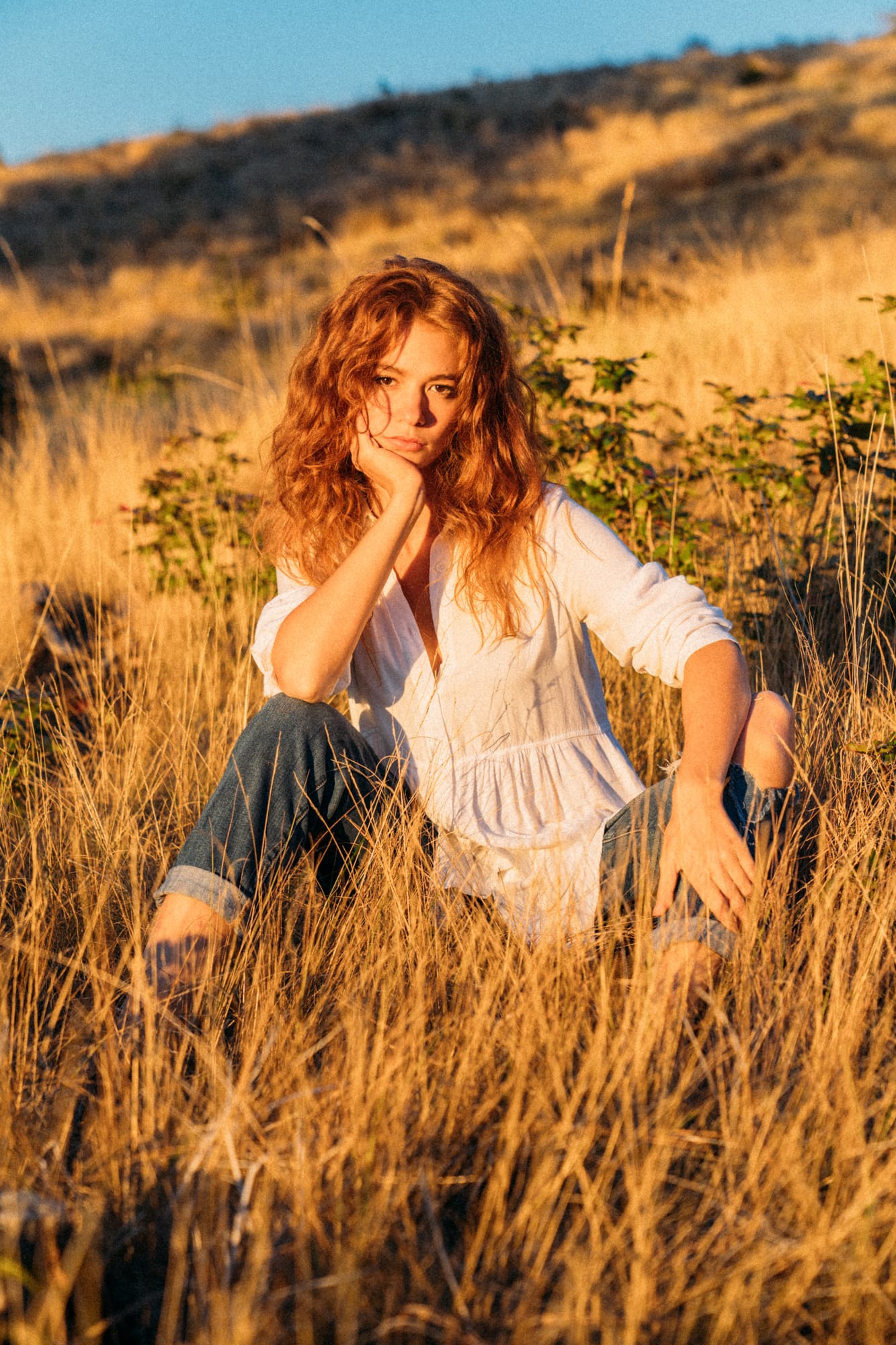 Mickie portraits story frame 10: A Kelowna portrait session for Mickie moving from golden grass and warm hillside light into lake-edge rocks, wind, white shirt styling and close editorial portraits.