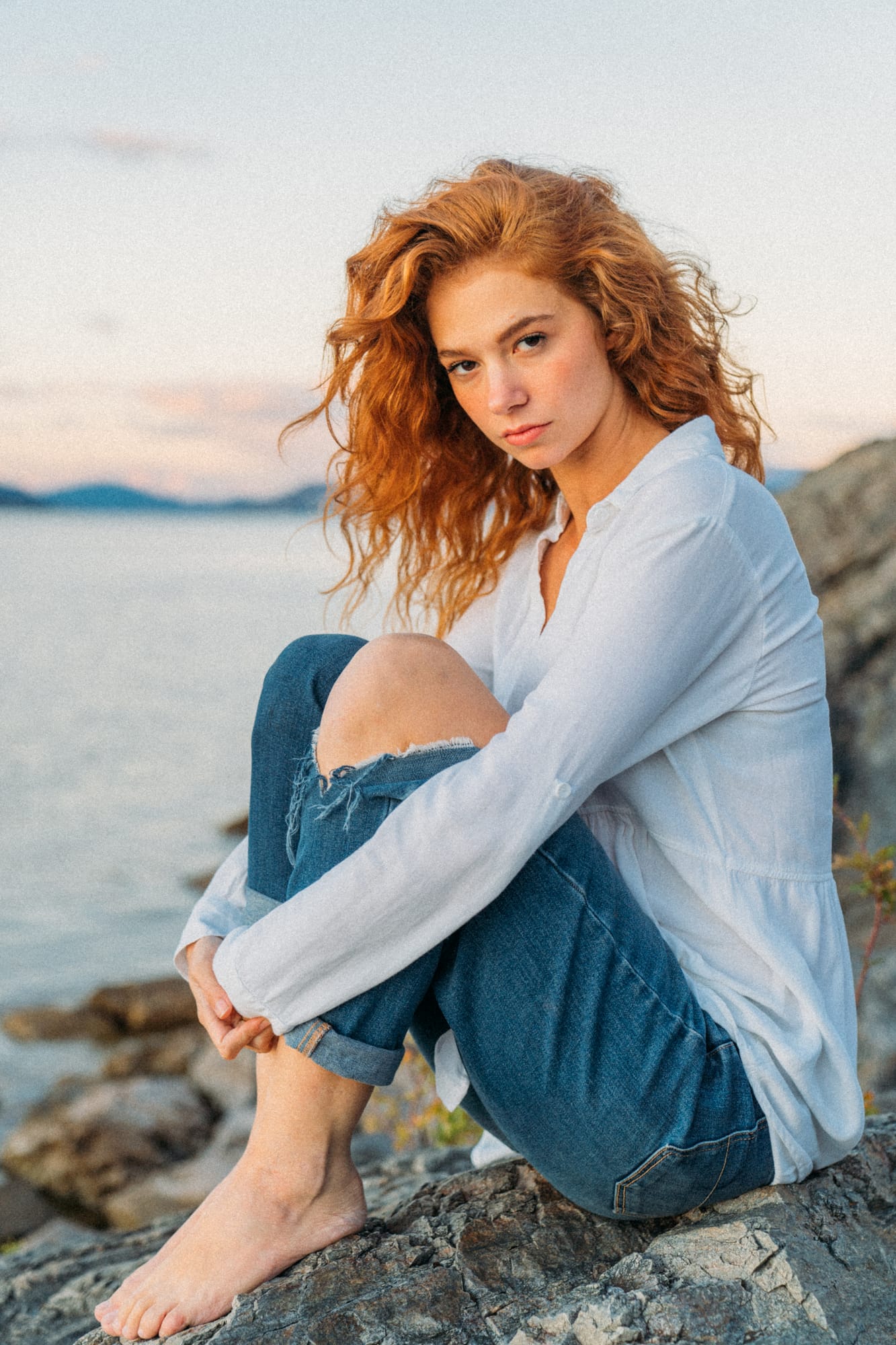 Mickie portraits story frame 17: A Kelowna portrait session for Mickie moving from golden grass and warm hillside light into lake-edge rocks, wind, white shirt styling and close editorial portraits.