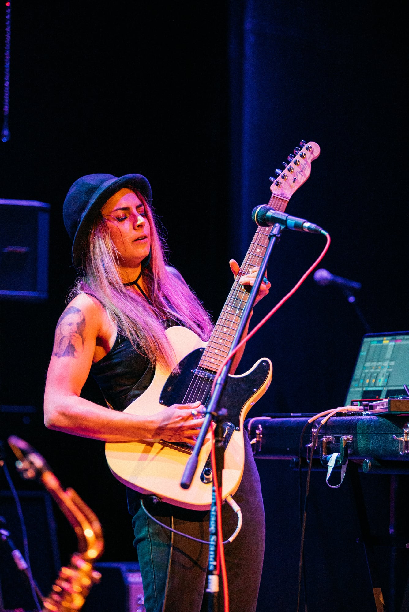 Woman with electric guitar and keyboard gear under blue and magenta stage light.