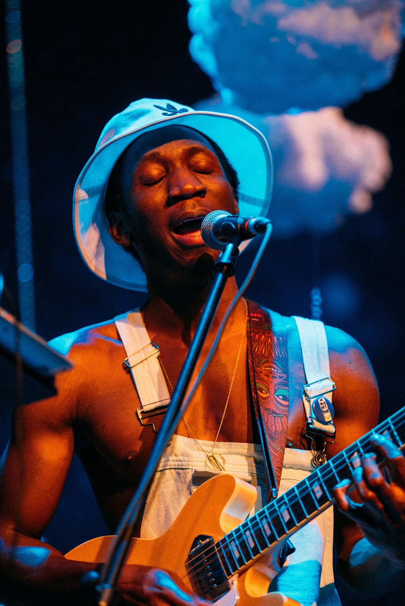 Close singer-guitarist portrait with blue hat and cloud backdrop.