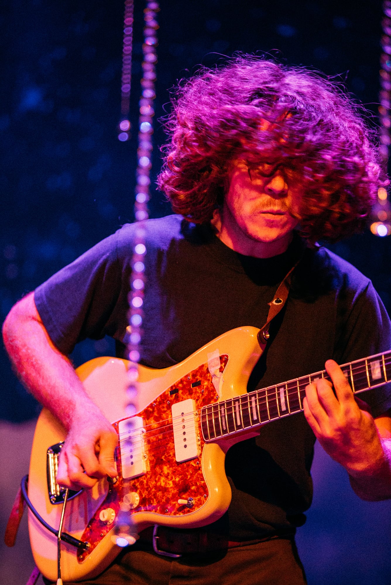 Curly-haired guitarist playing under magenta light.