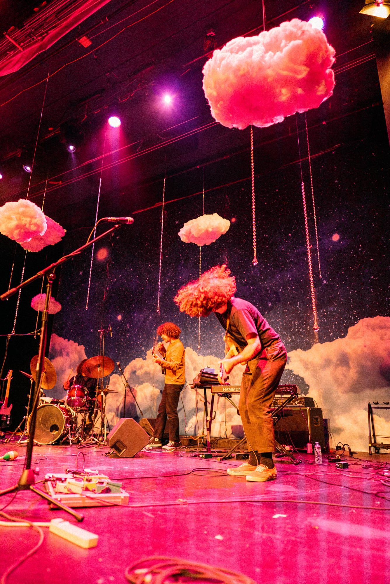 Band performing beneath hanging cloud props and red stage wash.