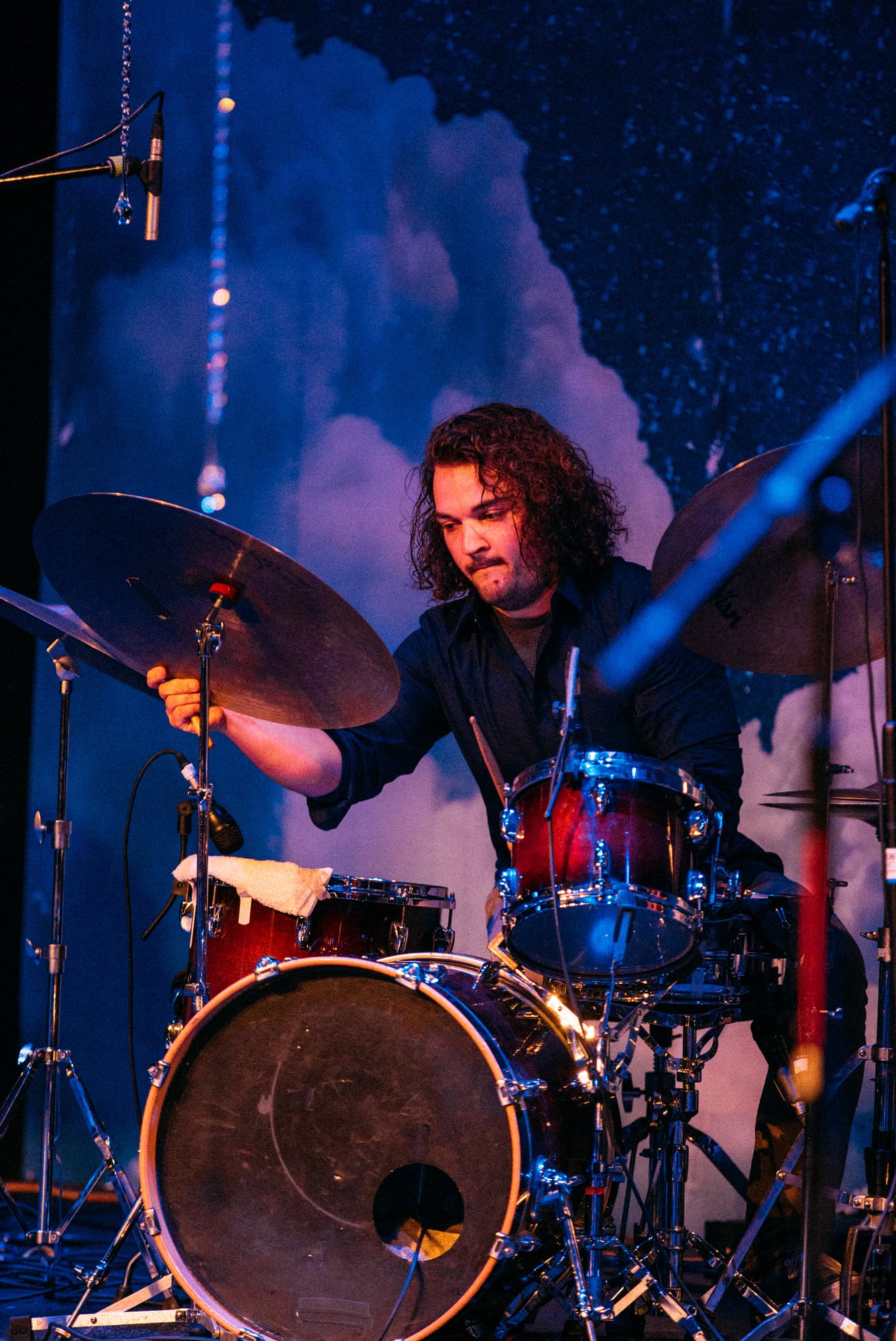 Drummer framed behind cymbals under blue stage light.