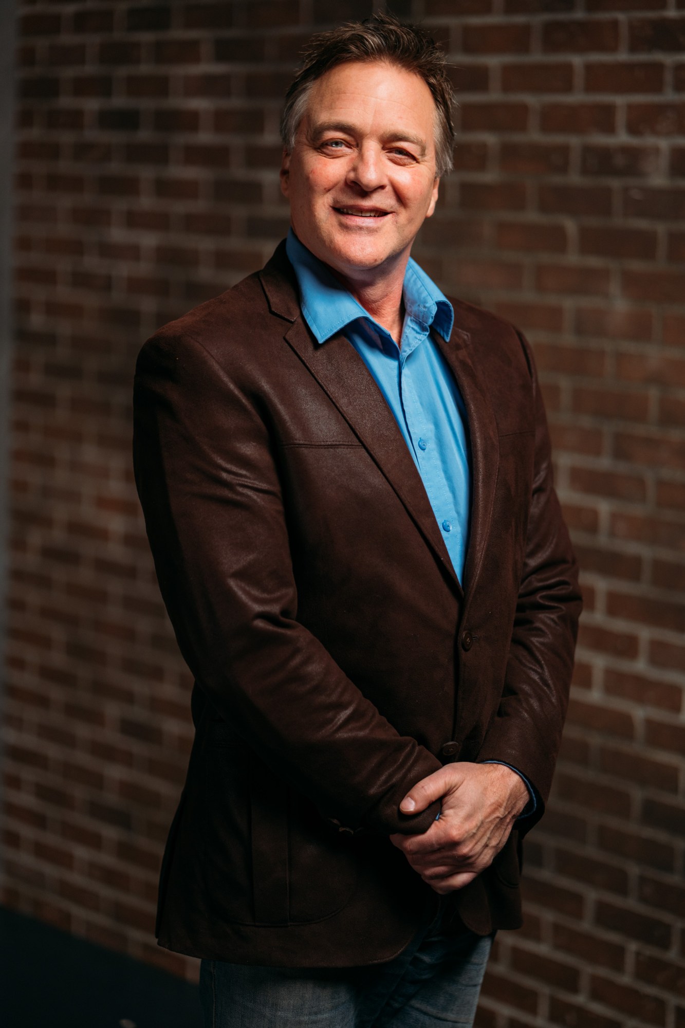Roark standing in a brown jacket with his hands together