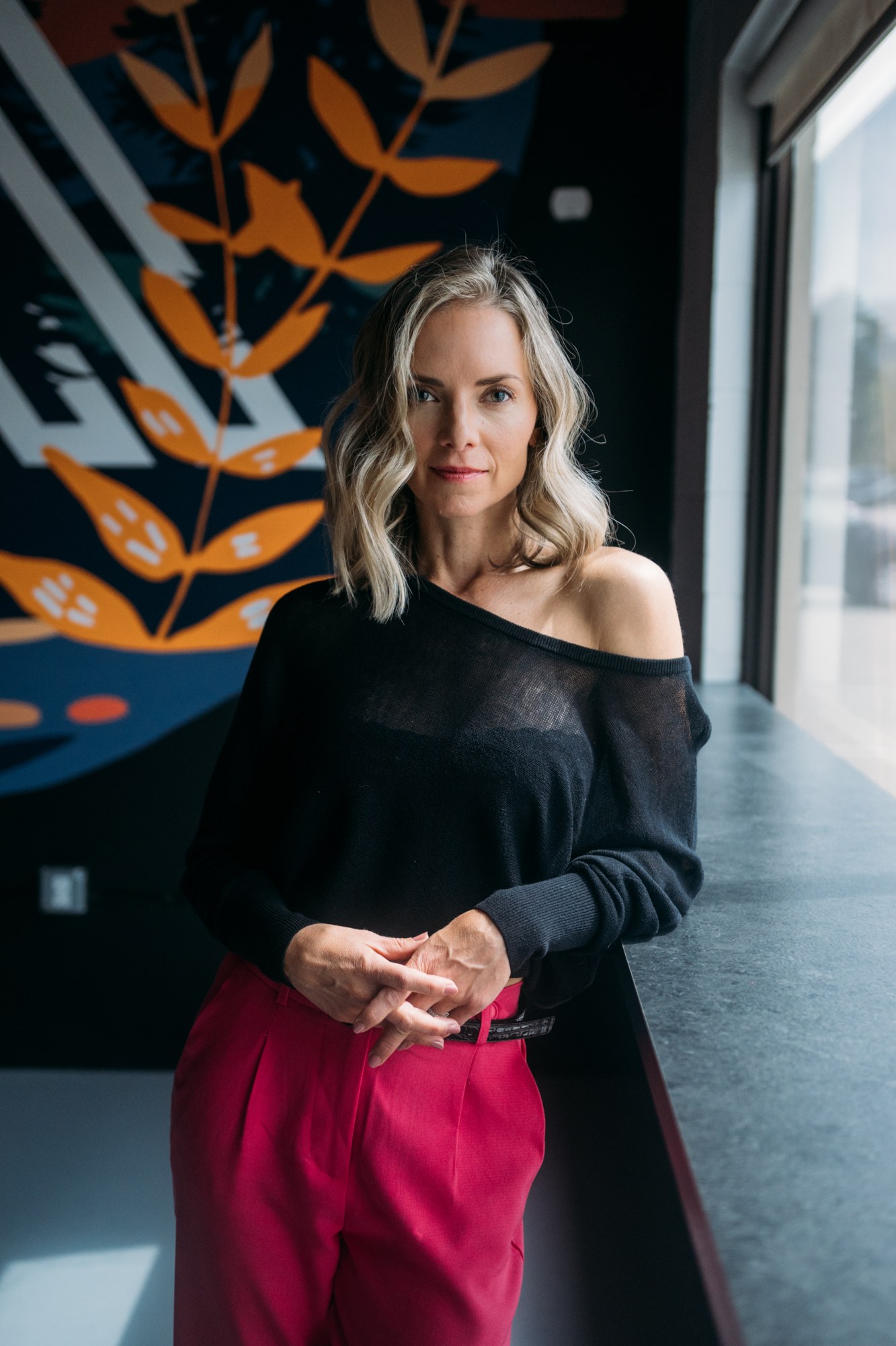 Sarah standing beside studio windows in pink trousers