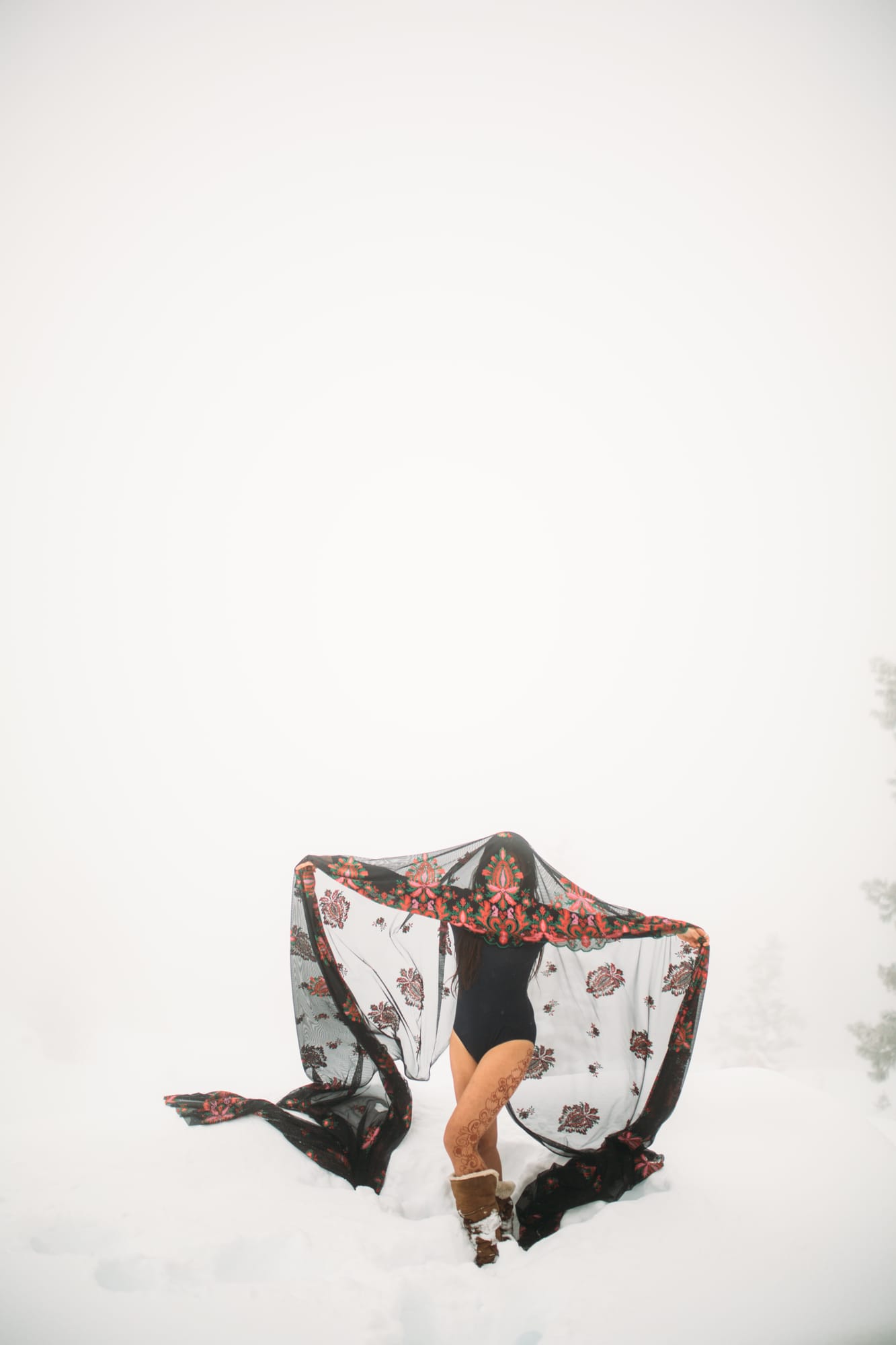 Sarah in the Snow portraits story frame 10: A winter portrait session for Sarah in Kelowna, with snow, black wardrobe, sheer floral fabric, whiteout atmosphere and bold movement in a cold landscape.