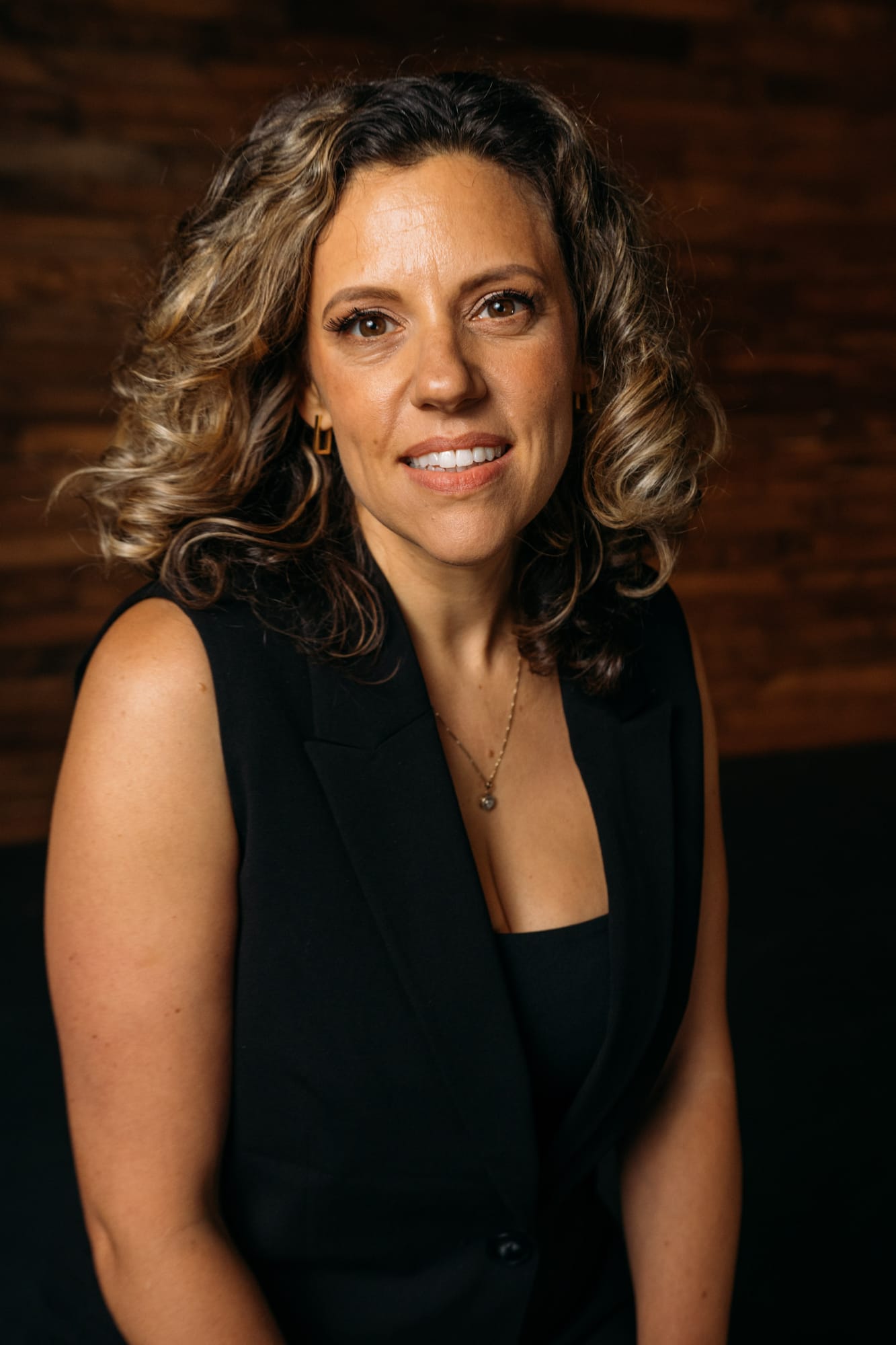 Trisha Miltimore corporate story frame 9: Personal-brand portraits for Trisha Miltimore in Kelowna, moving from clean business profile images into darker wood-wall portraits with black wardrobe and confident brand range..